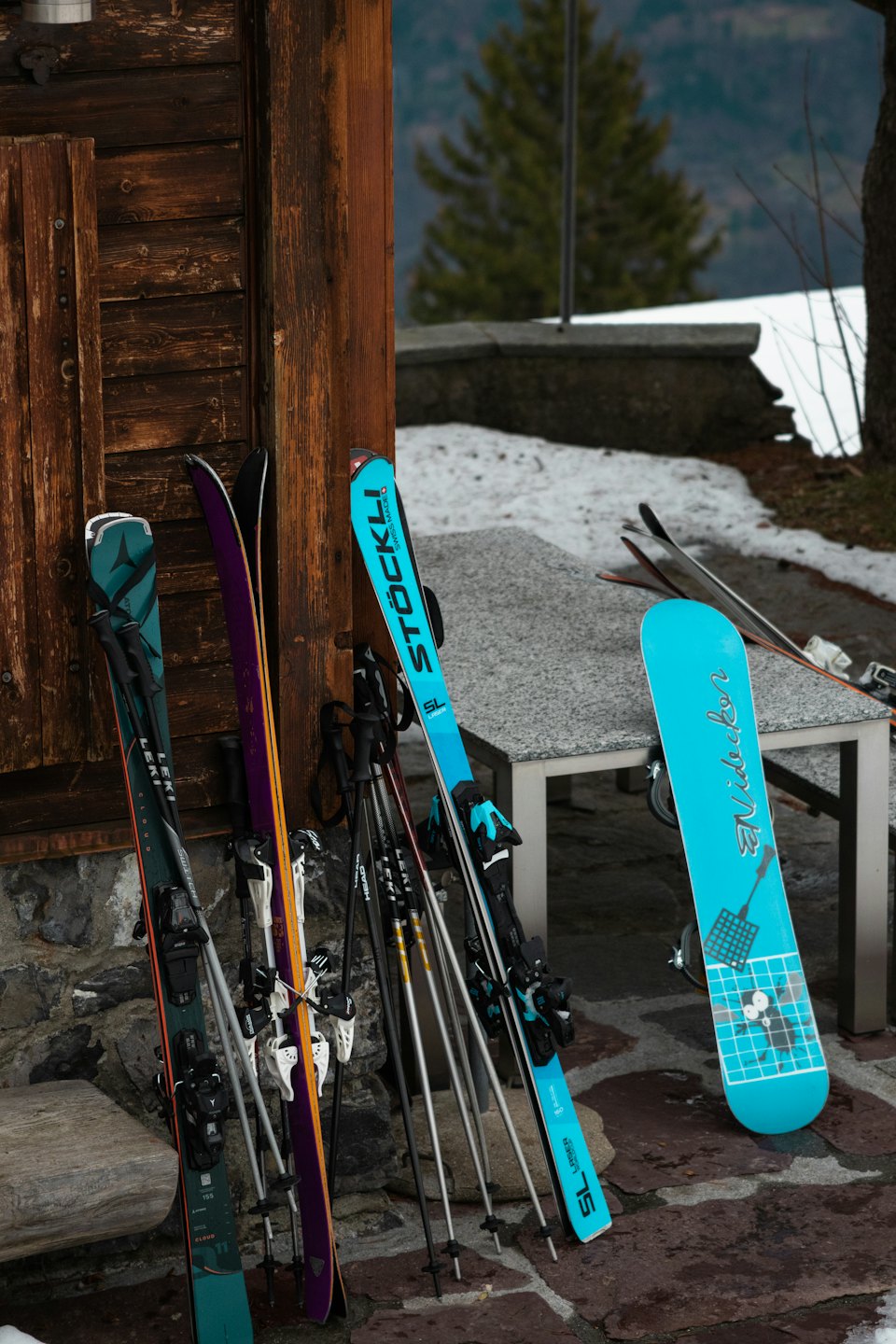 Sci, bastoncini e uno snowboard appoggiati a una parete di legno rustico.